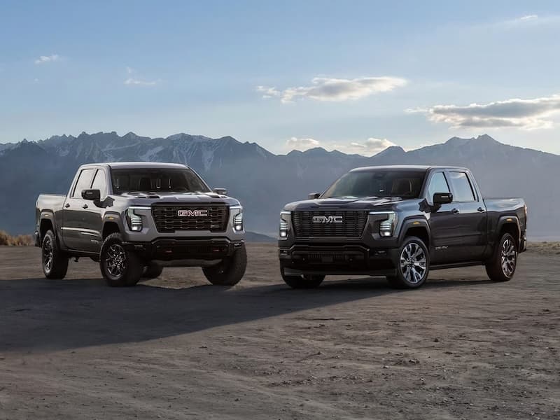 A tan 2026 GMC Sierra and a black 2026 GMC Sierra parked next to each other in front of the mountains.