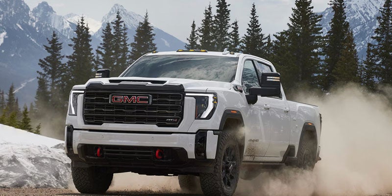 White GMC Sierra AT4X truck kicking up dust on a dirt road with snow-capped mountains and pine trees in background.